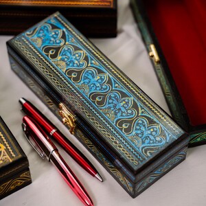 May include: An ornate, rectangular wooden pen box with a blue and gold floral design on the lid. Two red pens rest beside the box. Other decorative boxes are visible in the background, suggesting a collection of writing accessories.