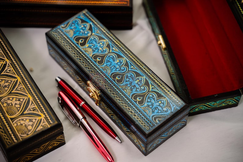 May include: An ornate, rectangular wooden pen box with a blue and gold floral design on the lid. Two red pens rest beside the box. Other decorative boxes are visible in the background, suggesting a collection of writing accessories.