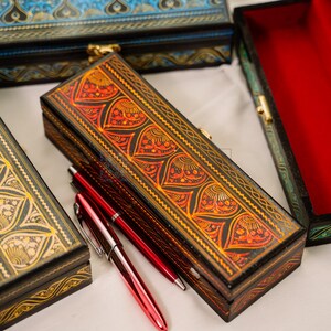 May include: Several decorative wooden boxes with intricate patterns and gold-colored clasps. One box is open, revealing a red interior. Two red pens are placed next to a box with an orange and black design.