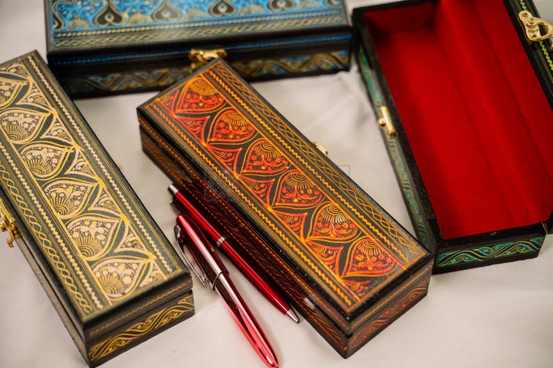 May include: Several decorative wooden boxes with intricate patterns and gold-colored clasps. One box is open, revealing a red interior. Two red pens are placed next to a box with an orange and black design.