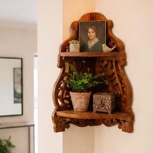 May include: A wooden corner shelf with intricate carvings, holding a small portrait, a potted plant, a decorative bird, and a small decorative box. The shelf is stained a warm brown color and is mounted on a neutral-colored wall.