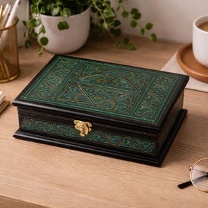 Könnte beinhalten: Eine rechteckige schwarze Holzbox mit einem verzierten teal- und goldfarbenen Design auf dem Deckel. Die Box hat einen goldenen Verschluss und steht auf einem Holzschreibtisch. Weitere Gegenstände sind vorhanden.