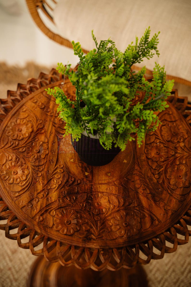 May include: A round, wooden side table with intricate floral carvings. The table is stained a warm brown and features a scalloped edge. A small potted plant with green foliage sits on top, adding a touch of nature.