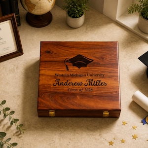 May include: A wooden box with a graduation cap design and the text "Western Michigan University, Andrew Miller, Class of 2026." A framed certificate, graduation cap, and diploma are also present. The box is approximately 10 inches wide.