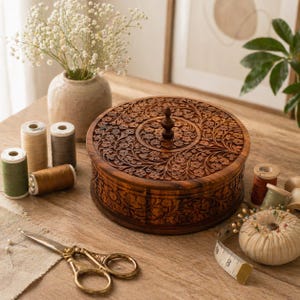 Puede incluir: Una caja redonda de madera con intrincadas tallas florales. La caja marrón tiene un pomo decorativo en la tapa. También hay carretes de hilo de varios colores, tijeras doradas y un alfiletero sobre la superficie de madera.