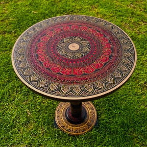 May include: A round, black wooden table with a hand-painted mandala design in red, gold, and black. The table has a single pedestal base.
