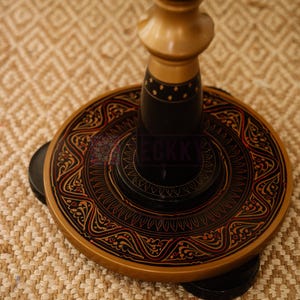 May include: A decorative wooden stand with a round base and ornate detailing. The base is black with red and gold accents, featuring intricate patterns. The stand's central column is light brown, and the overall design is traditional.