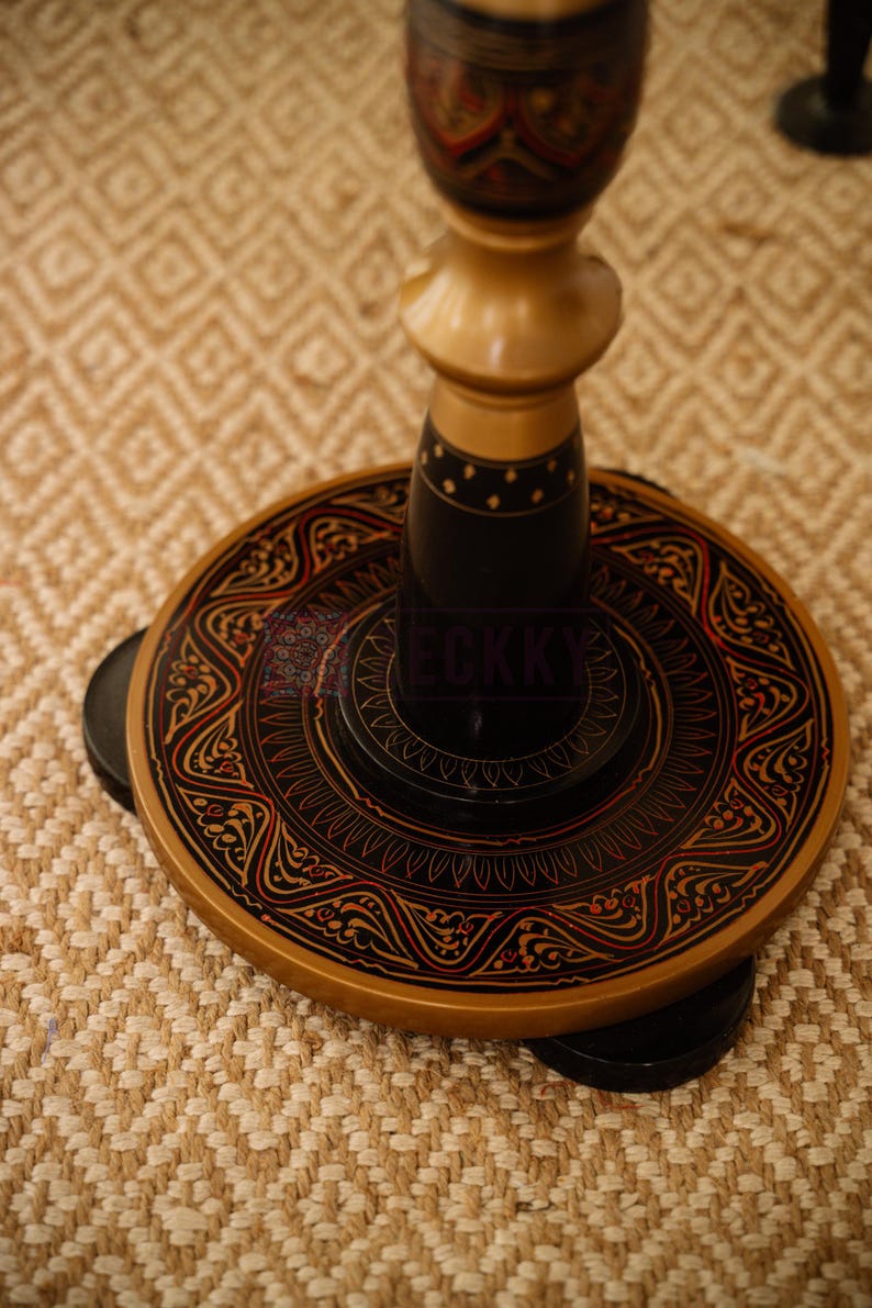 May include: A decorative wooden stand with a round base and ornate detailing. The base is black with red and gold accents, featuring intricate patterns. The stand's central column is light brown, and the overall design is traditional.