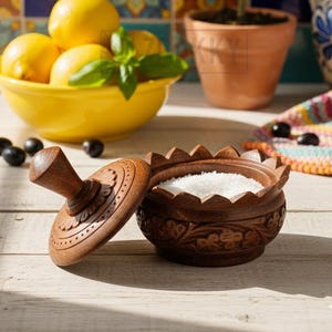 May include: A dark brown wooden salt cellar with a carved floral pattern and a scalloped rim, featuring a matching lid with a central handle. The container is filled with white salt, and the lid is placed beside it on a light wooden surface.