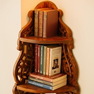 Puede incluir: Estantería esquinera de madera oscura con tres niveles, llena de libros de varios tamaños y colores. La estantería tiene detalles tallados decorativos. Los libros tienen lomos y cubiertas de diferentes colores, creando una exhibición visualmente atractiva.
