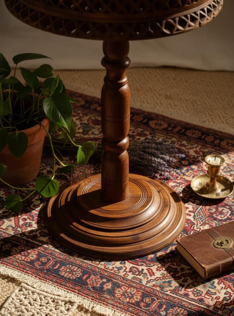 May include: A detailed shot of a dark brown wooden side table with a carved top and a turned pedestal base. The table is set on a patterned rug, with a potted plant, a candle holder, and a leather-bound book nearby. The table's height is approximately 28 inches.