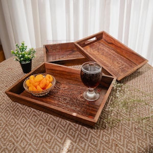 May include: Three intricately carved wooden serving trays. One tray holds a glass bowl of orange snacks and a glass of dark liquid. The trays are brown with floral and geometric patterns. The trays are on a patterned surface.