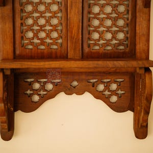 May include: A brown wooden wall shelf featuring two upper panels with intricate star-shaped lattice cutouts and a lower scalloped apron also decorated with matching star-shaped cutouts. The structure includes a flat horizontal shelf surface.