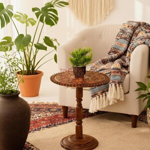 May include: A small, round, wooden side table with intricate carvings. The table has a dark brown finish and a pedestal base. A small potted plant sits on top of the table. A patterned throw blanket with tassels is draped over a cream-colored armchair.