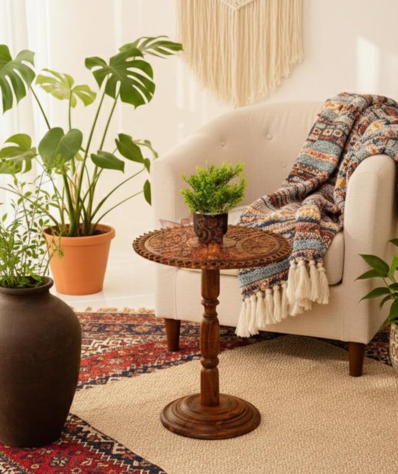 May include: A small, round, wooden side table with intricate carvings. The table has a dark brown finish and a pedestal base. A small potted plant sits on top of the table. A patterned throw blanket with tassels is draped over a cream-colored armchair.