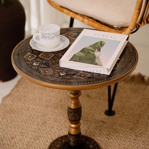 May include: A round, ornate side table with a gold-colored rim and intricate dark brown patterns. A white teacup and saucer with blue floral designs sit on the table, along with a book titled "CEREAL". The table is approximately 24 inches in diameter.