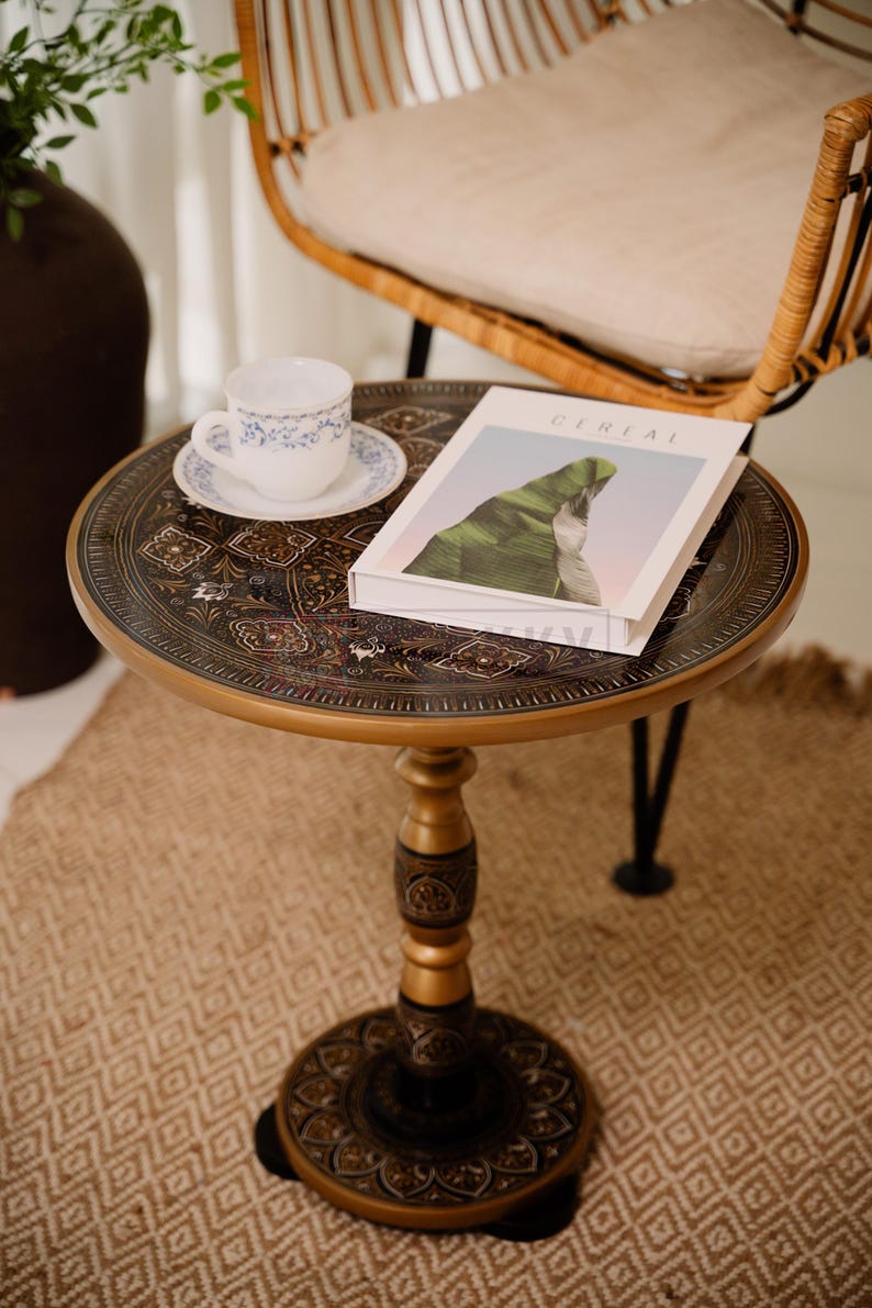 May include: A round, ornate side table with a gold-colored rim and intricate dark brown patterns. A white teacup and saucer with blue floral designs sit on the table, along with a book titled "CEREAL". The table is approximately 24 inches in diameter.