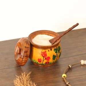 May include: A wooden salt cellar with a lid and a small wooden spoon. The cellar is painted with a yellow background and floral designs in red, green, and yellow. The cellar is filled with white salt.