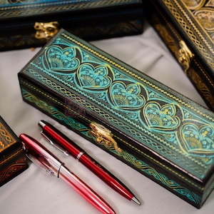 May include: Several decorative boxes with intricate designs and gold clasps are displayed. The boxes feature heart and floral patterns in teal, gold, and red. Two red pens are placed near the boxes, suggesting a stationery theme.