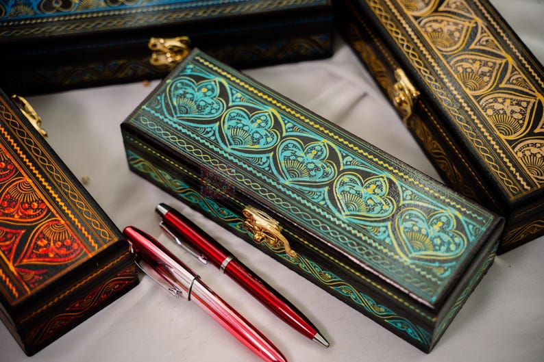 May include: Several decorative boxes with intricate designs and gold clasps are displayed. The boxes feature heart and floral patterns in teal, gold, and red. Two red pens are placed near the boxes, suggesting a stationery theme.