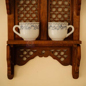 May include: A brown wooden wall shelf with intricate lattice cutouts and two white ceramic cups featuring a delicate blue floral pattern. The shelf has a flat horizontal surface for display and decorative side supports with carved details.