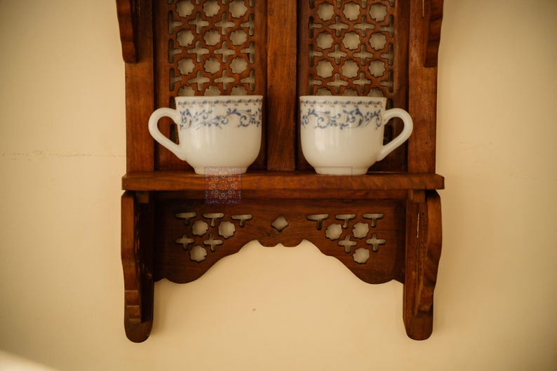 May include: A brown wooden wall shelf with intricate lattice cutouts and two white ceramic cups featuring a delicate blue floral pattern. The shelf has a flat horizontal surface for display and decorative side supports with carved details.