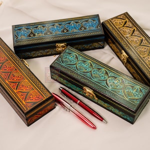 May include: Four rectangular, decorative boxes with intricate, embossed designs in shades of red, blue, green, and gold. Each box has a hinged lid and a gold-colored clasp. Two red pens are placed near one of the boxes.