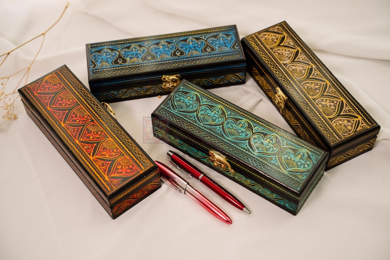 May include: Four rectangular, decorative boxes with intricate, embossed designs in shades of red, blue, green, and gold. Each box has a hinged lid and a gold-colored clasp. Two red pens are placed near one of the boxes.