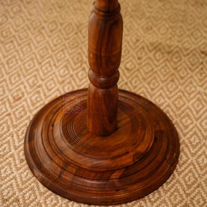May include: A close-up of a dark brown wooden table base and pedestal. The base is round with concentric circular grooves. The pedestal is a turned design with a bulbous center. The table is set on a beige patterned rug.