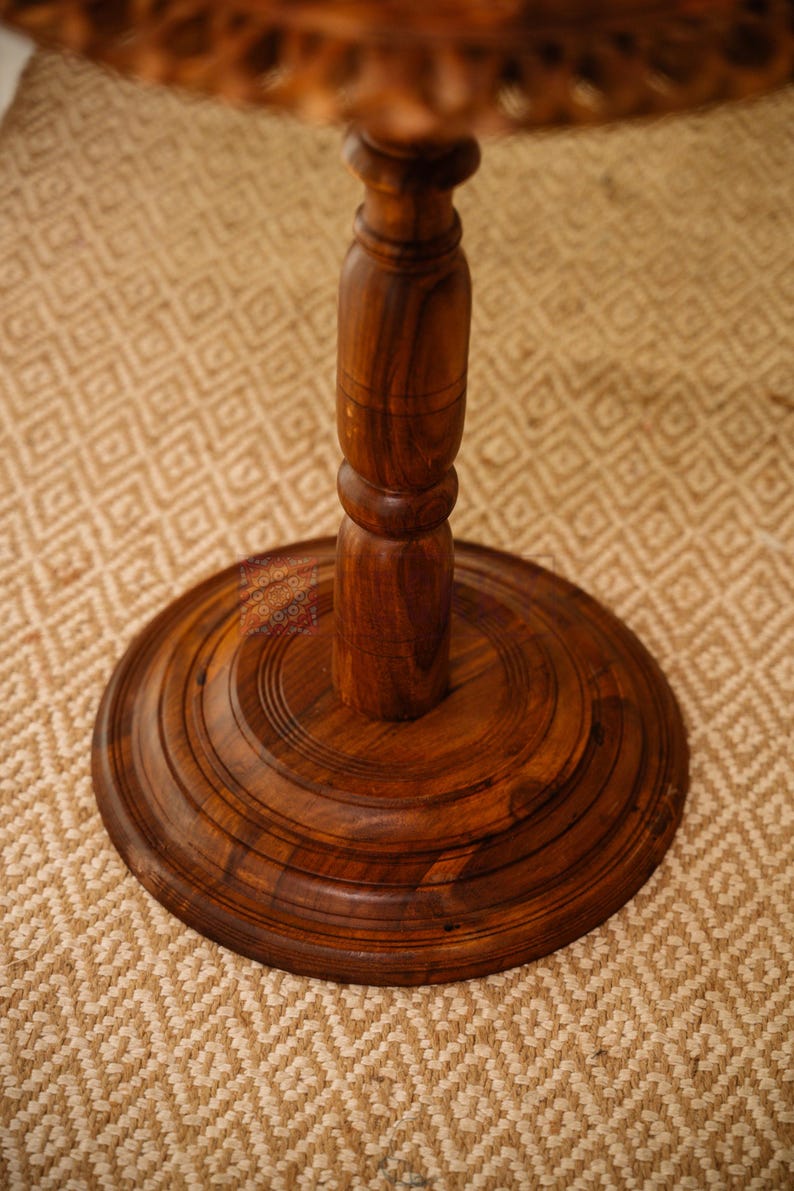 May include: A close-up of a dark brown wooden table base and pedestal. The base is round with concentric circular grooves. The pedestal is a turned design with a bulbous center. The table is set on a beige patterned rug.