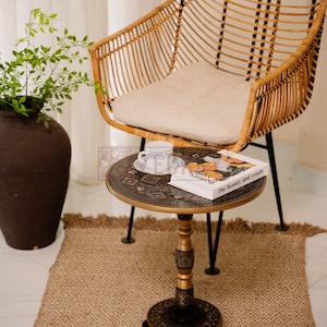 May include: A rattan chair with a cream-colored cushion sits near a small, round table. The table has a decorative top with a book titled "The beauty and youth" and a teacup. A woven rug and a large brown vase with greenery complete the scene.