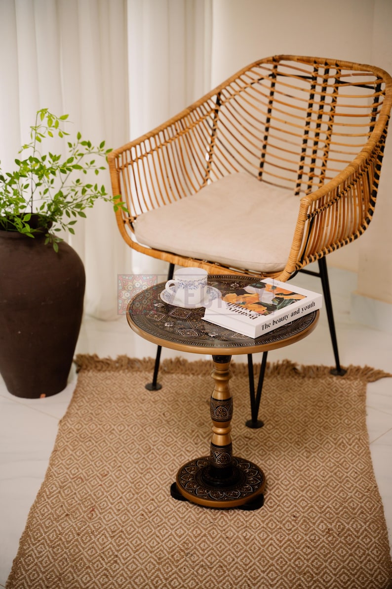 May include: A rattan chair with a cream-colored cushion sits near a small, round table. The table has a decorative top with a book titled "The beauty and youth" and a teacup. A woven rug and a large brown vase with greenery complete the scene.