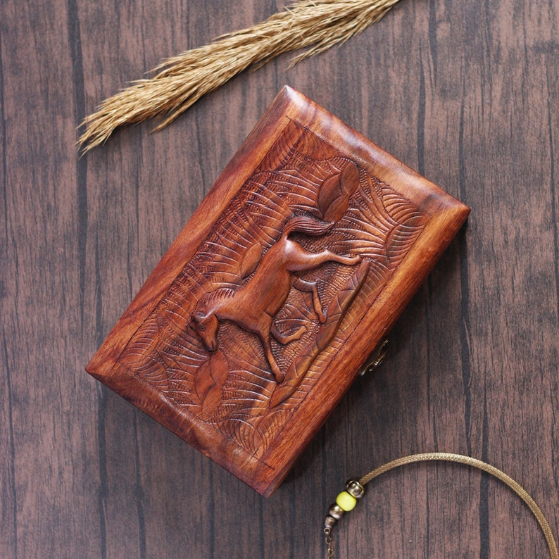 May include: A wooden box with a carved horse design. The box is made of dark brown wood and has a hinged lid. The horse is carved in relief and is facing to the right. The box is sitting on a wooden surface.