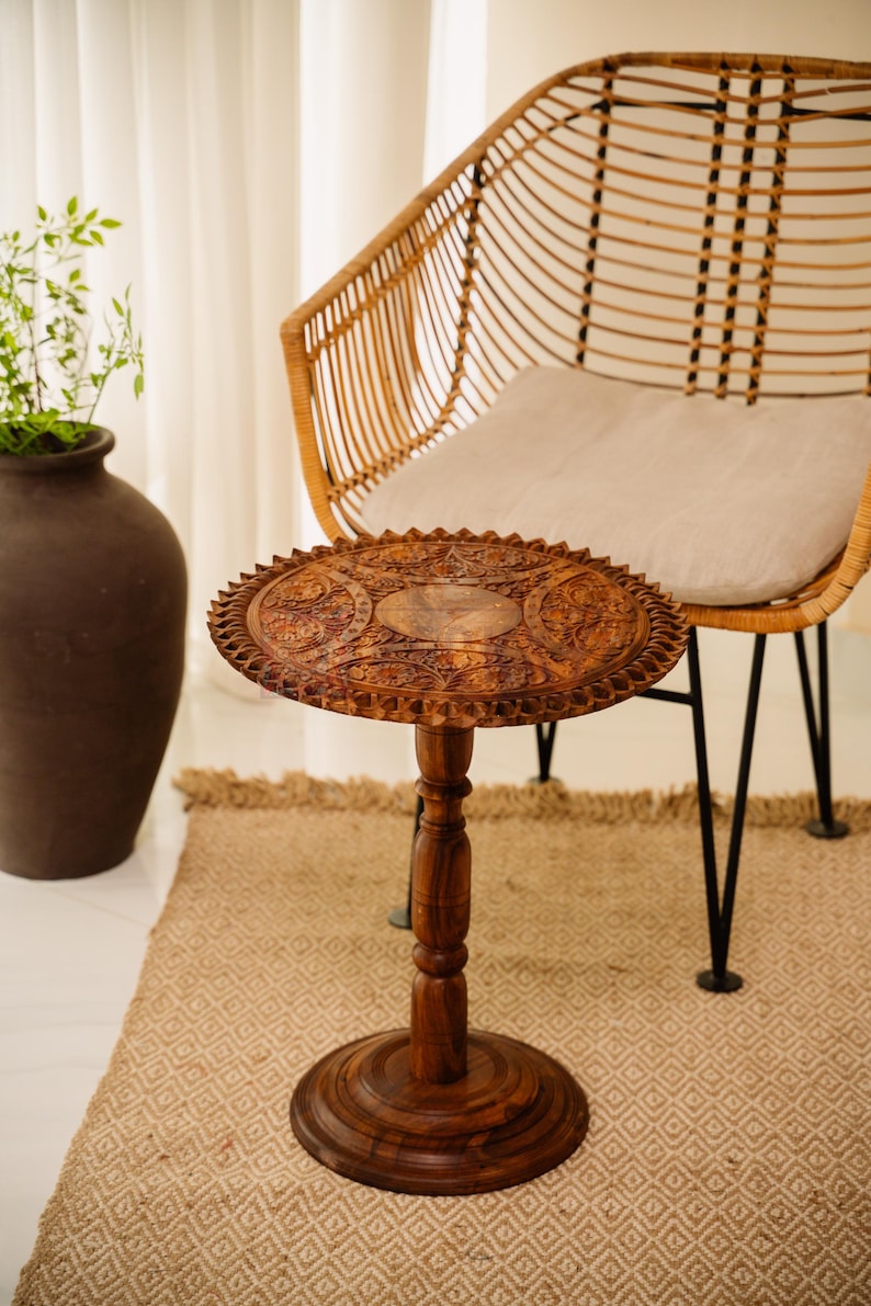 May include: A small, round, wooden side table with intricate carved floral patterns. The table has a dark brown finish and a single pedestal base. It is placed on a patterned rug, near a woven chair and a large brown vase.