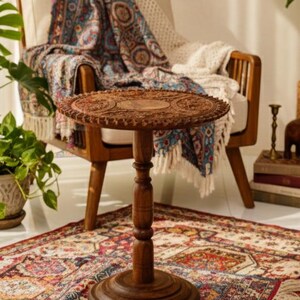 May include: A small, round, wooden side table with intricate carved details on the tabletop. The table has a dark brown finish and stands on a pedestal base. It is placed on a patterned rug near a chair with a blanket.