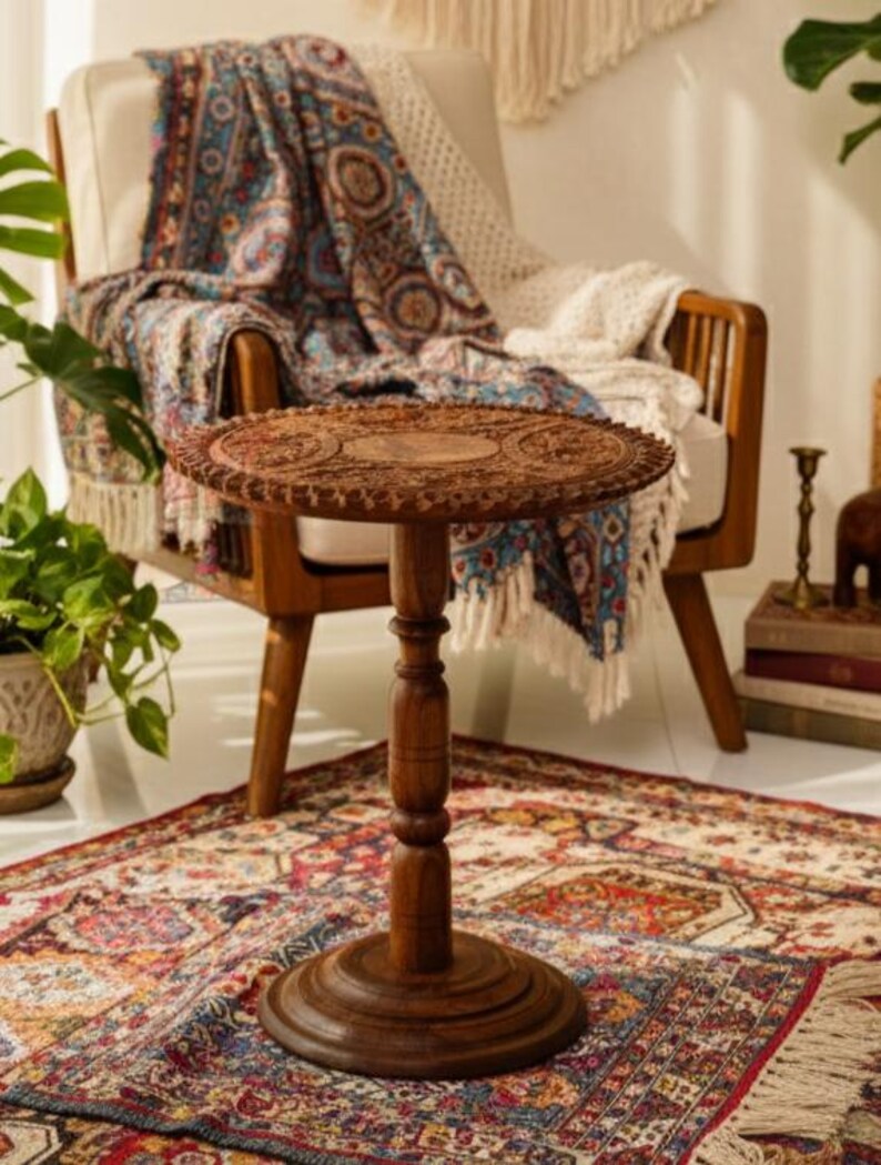 May include: A small, round, wooden side table with intricate carved details on the tabletop. The table has a dark brown finish and stands on a pedestal base. It is placed on a patterned rug near a chair with a blanket.