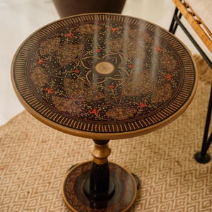 May include: A round, decorative side table with a black top featuring an intricate floral design in red and gold. The table has a gold-colored rim and base, and a black pedestal. The table is approximately 20 inches in diameter.