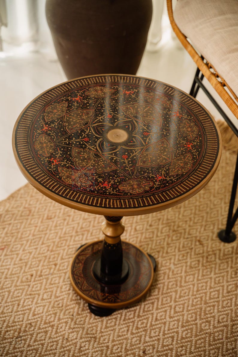 May include: A round, decorative side table with a black top featuring an intricate floral design in red and gold. The table has a gold-colored rim and base, and a black pedestal. The table is approximately 20 inches in diameter.