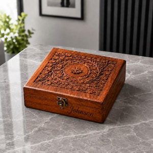 May include: A square, wooden box with intricate floral carvings on the lid. The box is a warm brown color and has a brass clasp. The name "Johnson" is engraved on the front. The box is sitting on a marble surface.