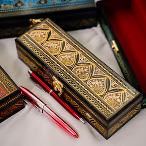 May include: Several decorative boxes with intricate gold and black designs are displayed. Two red pens are next to a rectangular box with a gold clasp. The boxes have a variety of colors, including red, black, blue, and green.