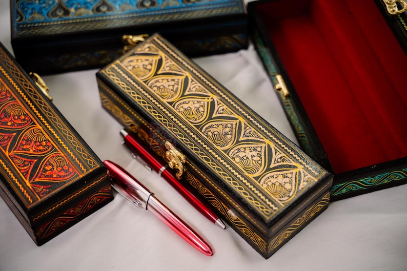 May include: Several decorative boxes with intricate gold and black designs are displayed. Two red pens are next to a rectangular box with a gold clasp. The boxes have a variety of colors, including red, black, blue, and green.