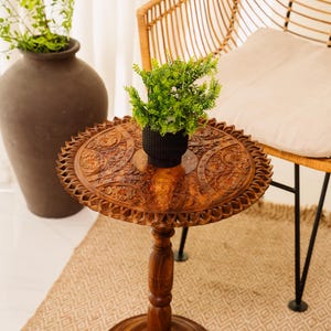 May include: A small, round, wooden side table with intricate carved floral patterns. The table has a dark brown finish and a single pedestal base. A small black pot with green plants sits on top. A woven chair is in the background.