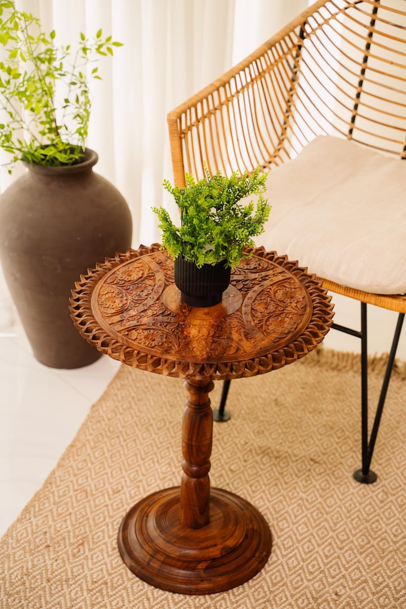 May include: A small, round, wooden side table with intricate carved floral patterns. The table has a dark brown finish and a single pedestal base. A small black pot with green plants sits on top. A woven chair is in the background.