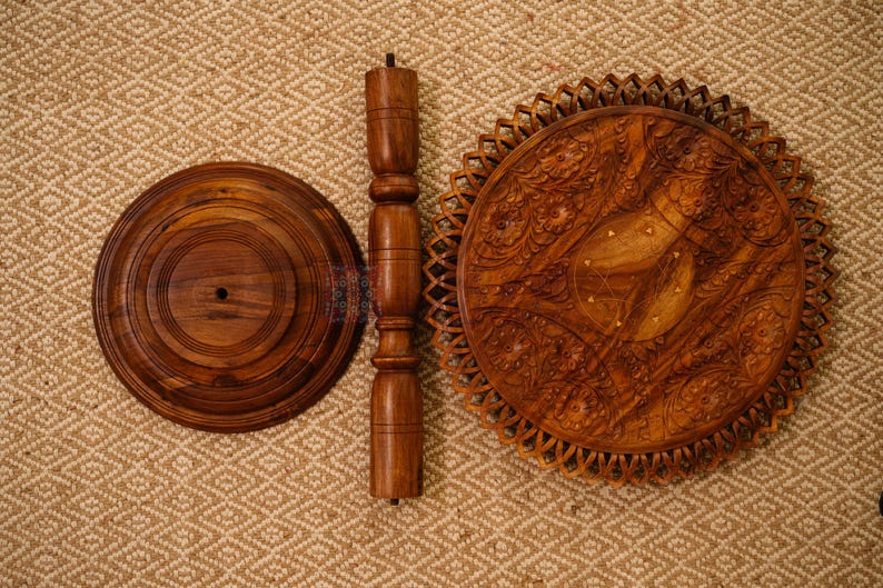May include: A disassembled wooden side table with a round base, a central support, and a decorative tabletop. The tabletop features intricate carved floral patterns. The wood has a warm, brown tone.