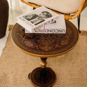 May include: A round, decorative side table with a detailed, ornate design on the tabletop. The table has a black and gold color scheme, with a central pedestal base. Two books are stacked on top of the table. The text "ACNE PAPER" is visible on the top book.
