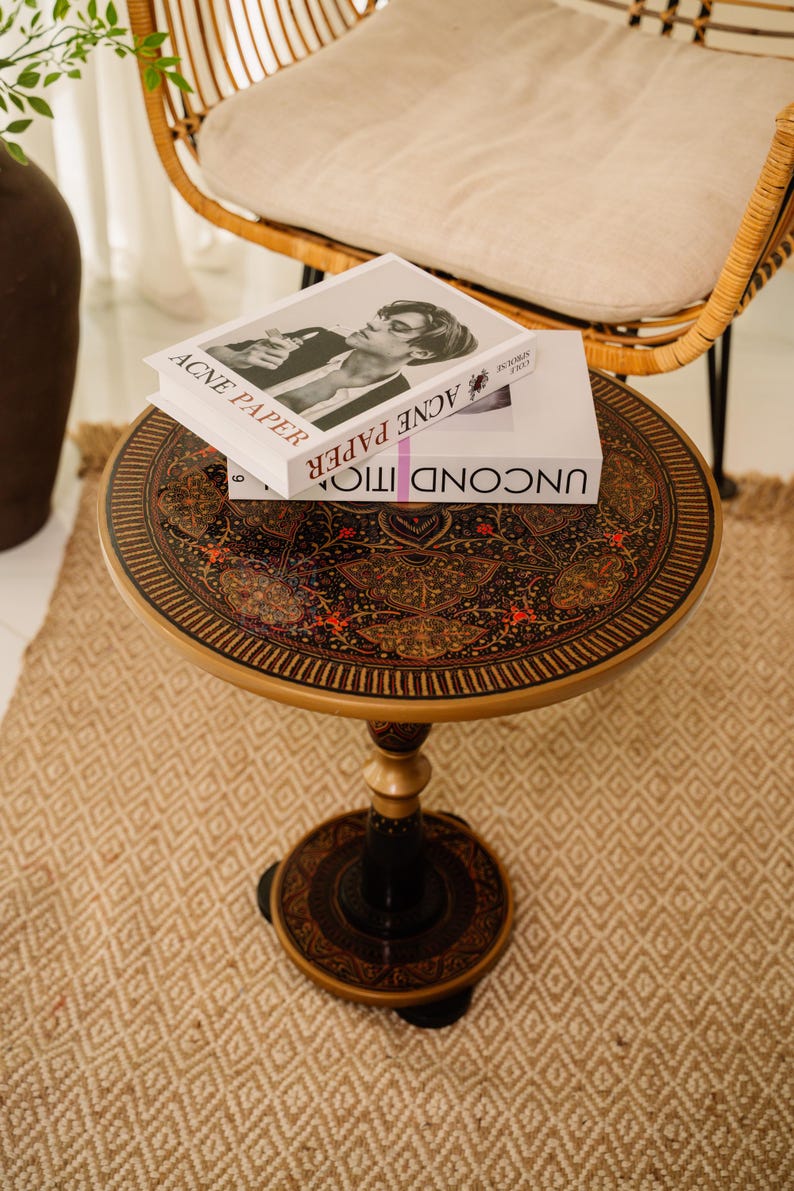 May include: A round, decorative side table with a detailed, ornate design on the tabletop. The table has a black and gold color scheme, with a central pedestal base. Two books are stacked on top of the table. The text "ACNE PAPER" is visible on the top book.