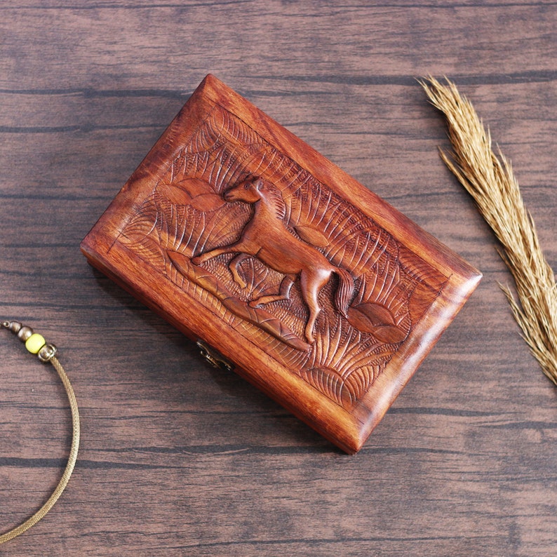 May include: A wooden box with a carved horse design. The box is made of dark brown wood and has a hinged lid. The horse is carved in relief and is facing to the right. The box is sitting on a wooden surface.