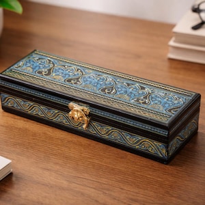May include: A rectangular wooden box with a hinged lid, featuring a blue and gold floral design. The box has a gold-colored clasp and is set on a wooden surface. Pens and a notepad are visible nearby.