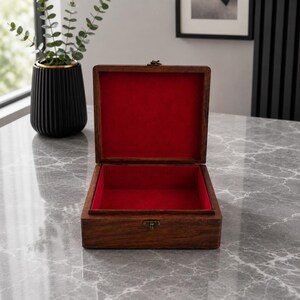 May include: An open wooden box with a red velvet interior. The box is rectangular with a hinged lid and a brass-colored clasp. The wood has a rich, dark brown color. The box is sitting on a marble-patterned surface.