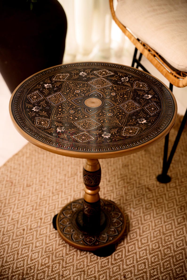 May include: A round side table with an ornate, dark brown and gold floral design on the tabletop. The table has a pedestal base and is set on a patterned rug. The table's design suggests an antique or vintage style.